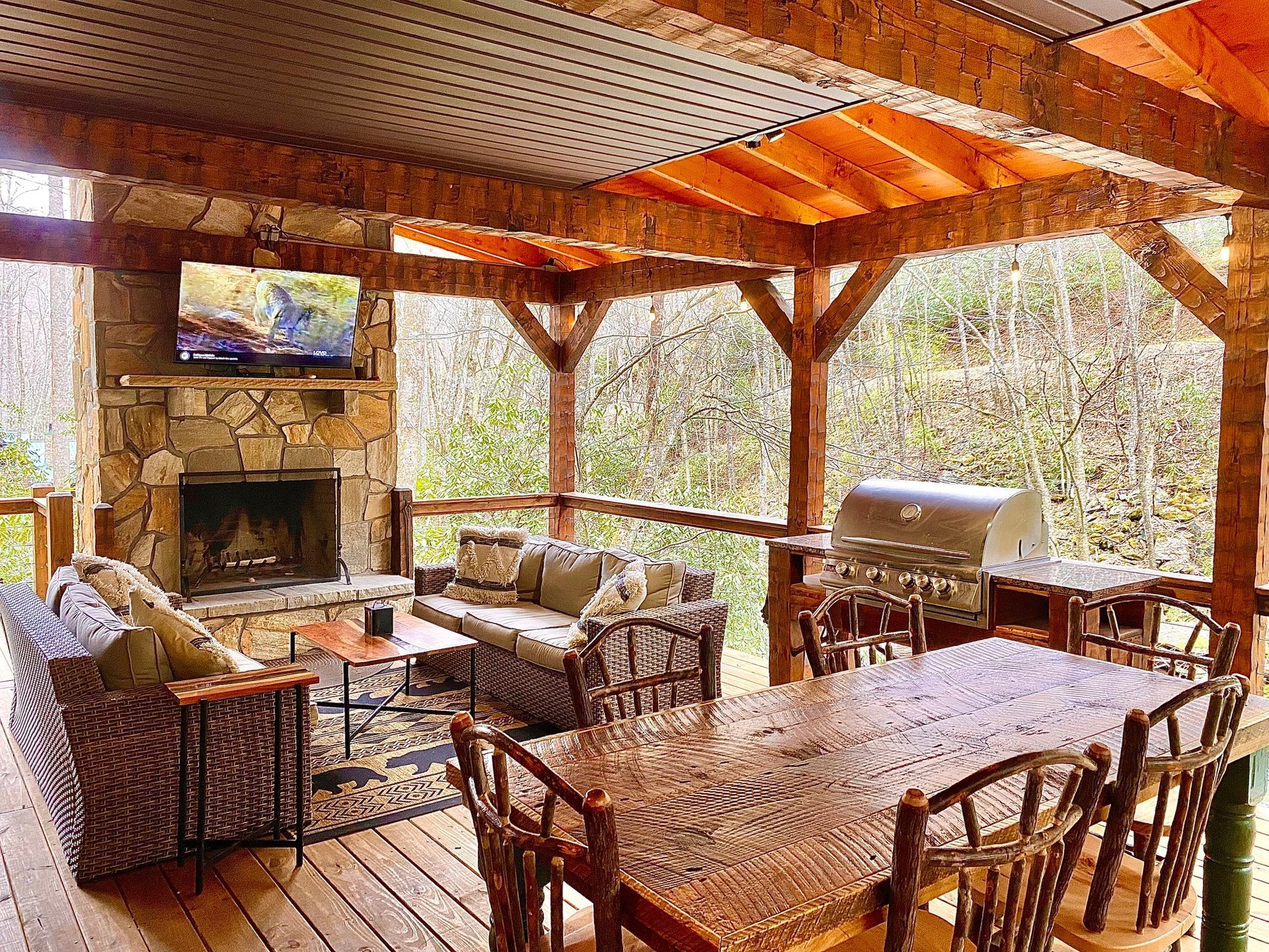 Covered outdoor deck with fireplace, seating, dining table, and grill in a wooded setting.