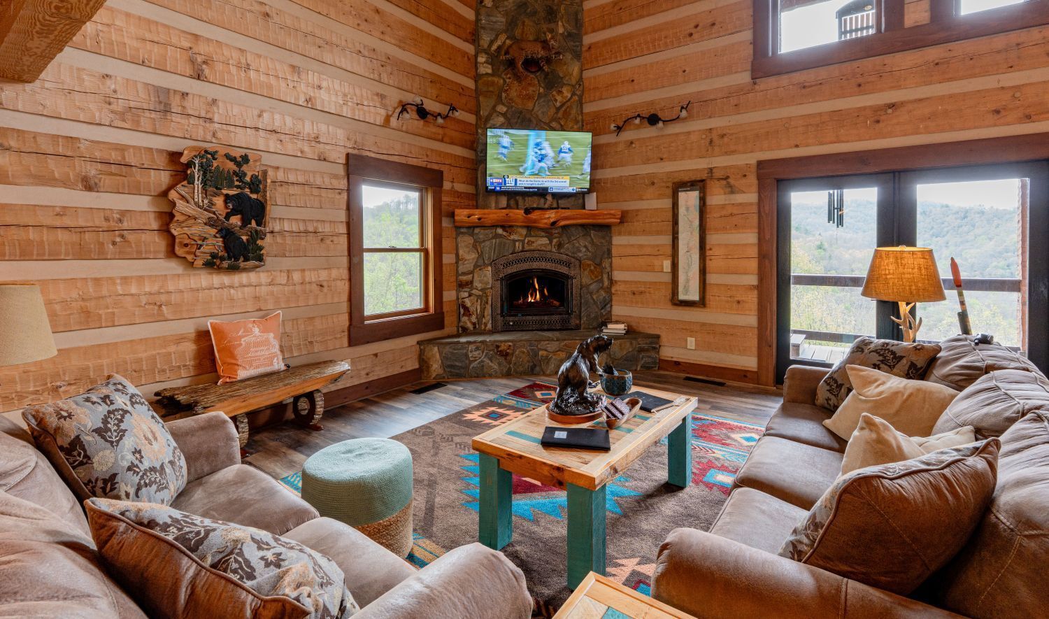 Cozy log cabin living room with stone fireplace, sectional sofa, windows, and TV.