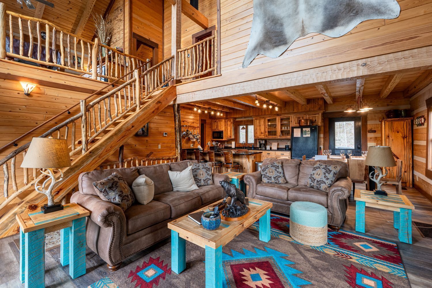 Living room in a log cabin with brown sofas, turquoise tables, a large rug, and a staircase.