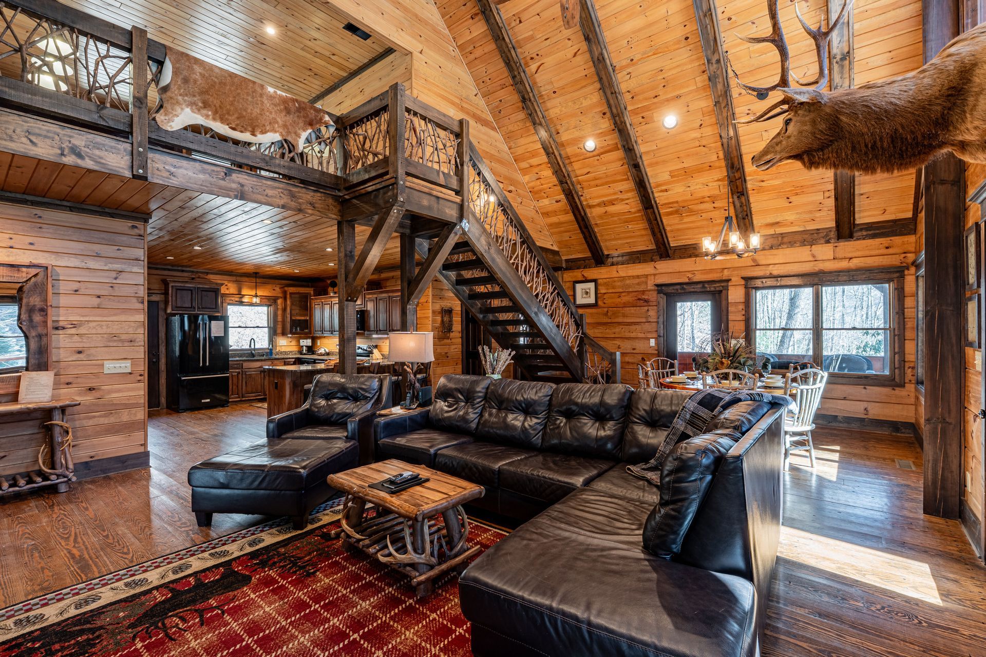 Rustic cabin interior with leather sectional, fireplace, staircase, and taxidermied deer heads.