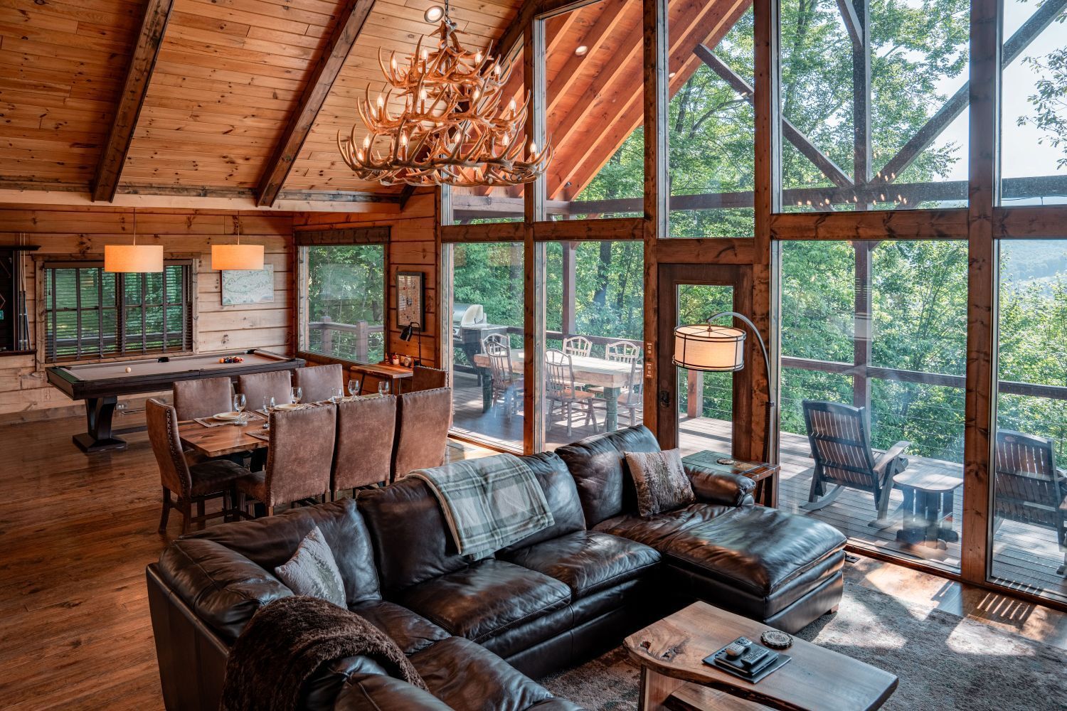 Luxe cabin interior: leather sectional, dining table, floor-to-ceiling windows with forest view, chandelier.