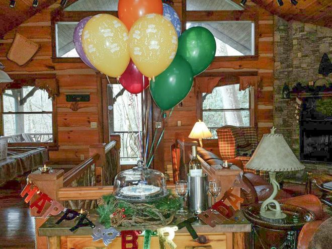 Balloons over a cake on a decorated table in a log cabin with a fireplace and windows.