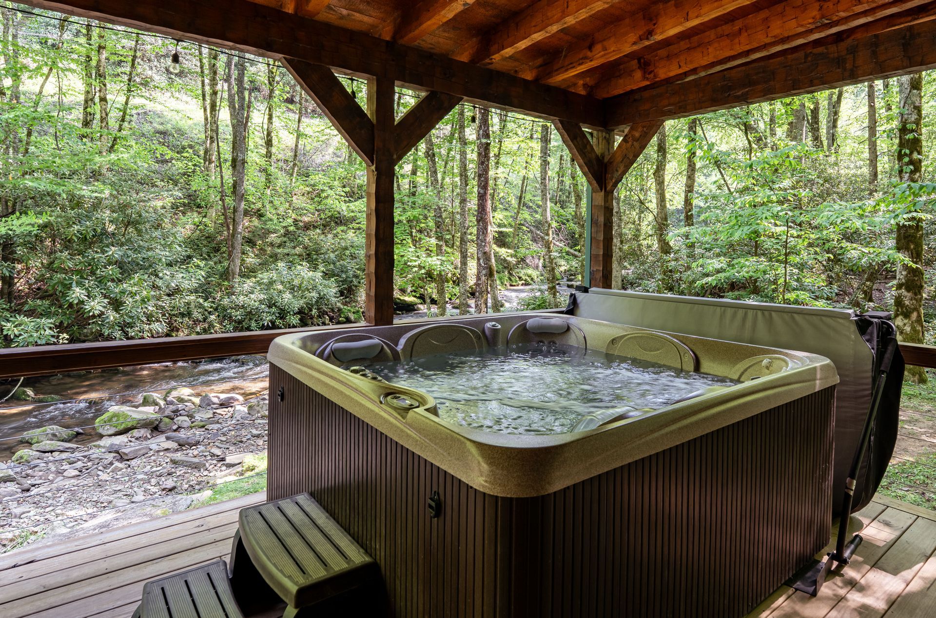 Cabin with porch and string lights next to a flowing river, surrounded by trees.