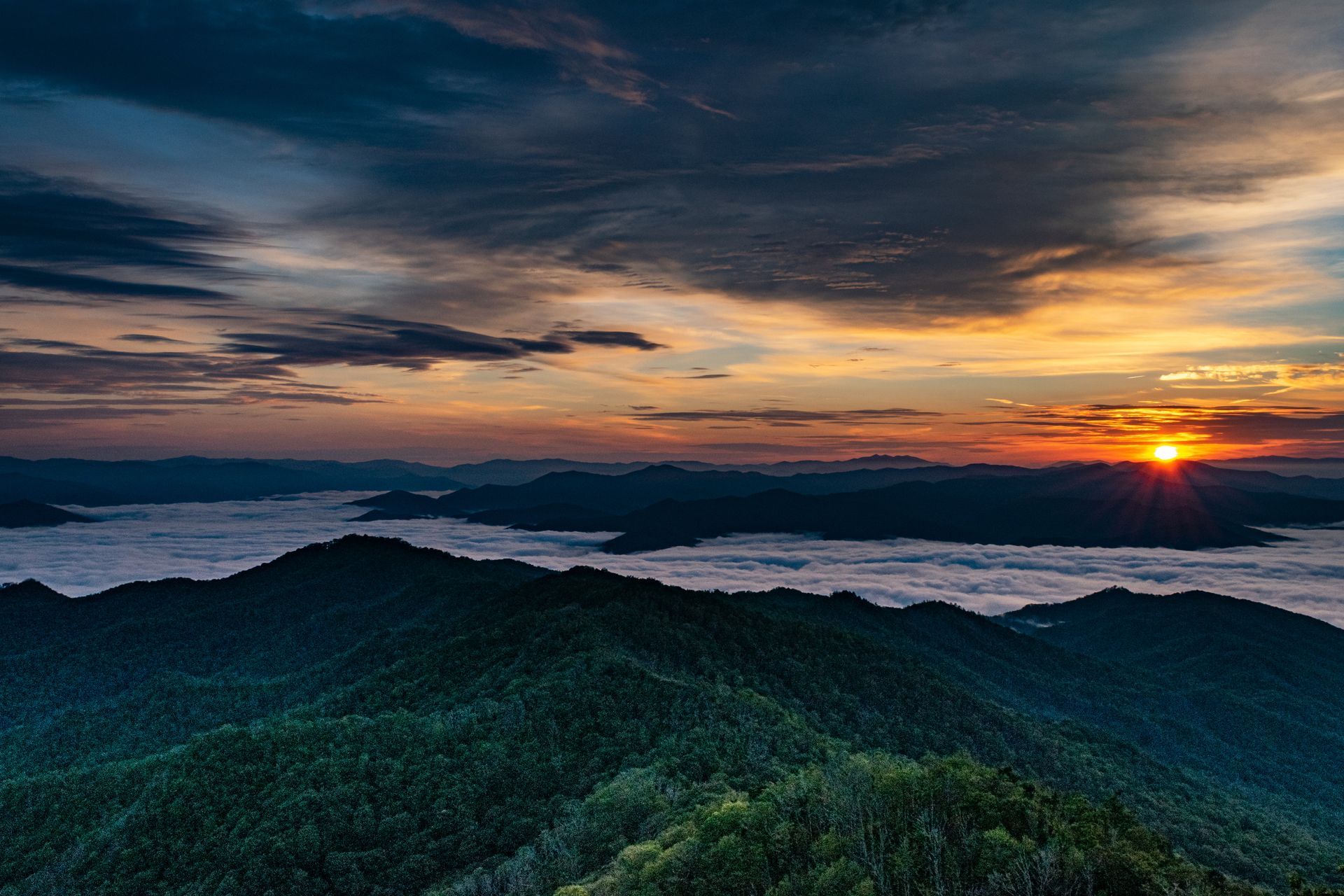 Sunrise over mountain range with low-lying fog. Golden light, dark green hills, and cloudy sky.