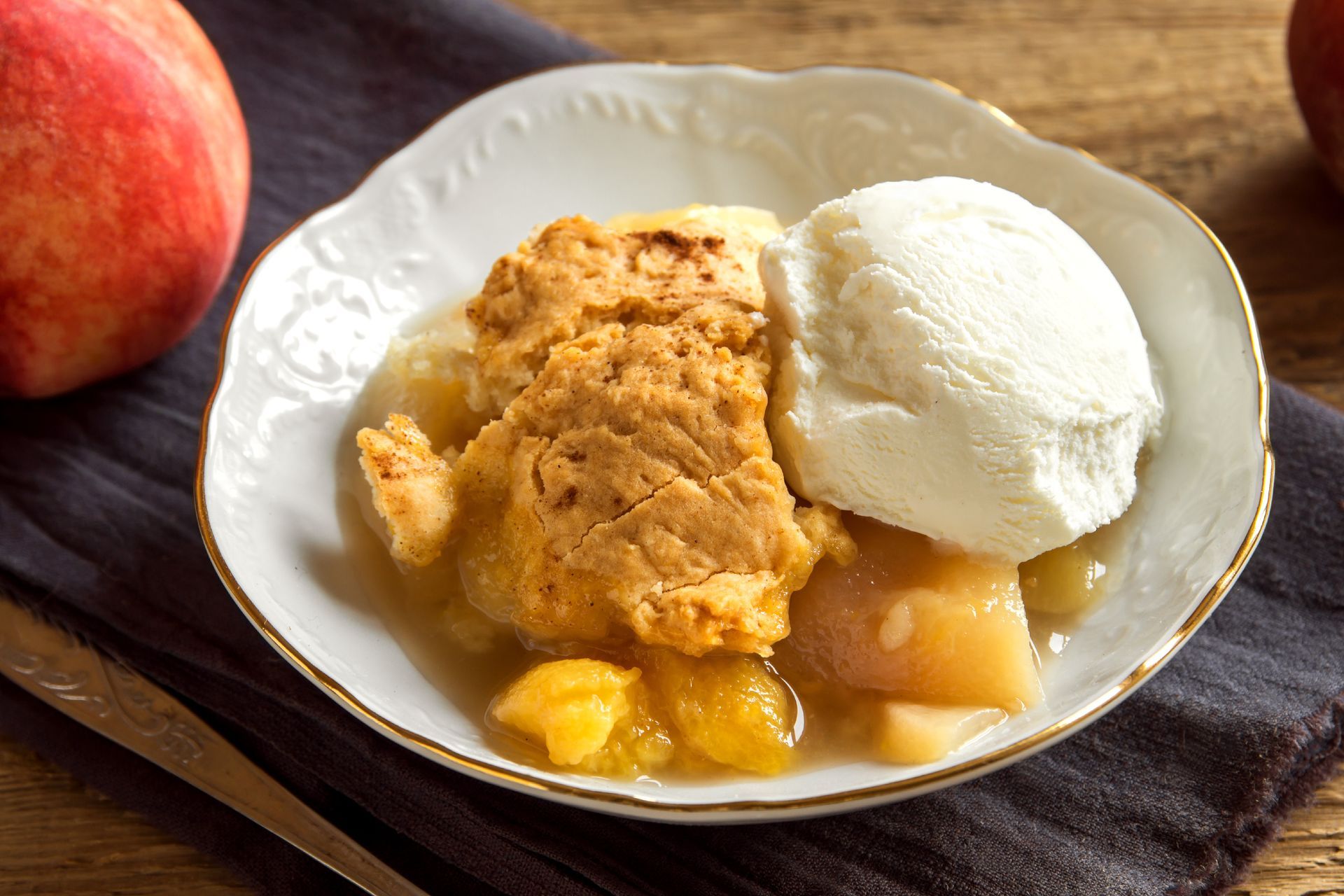 Peach cobbler with vanilla ice cream in a white ornate bowl on a wooden table.