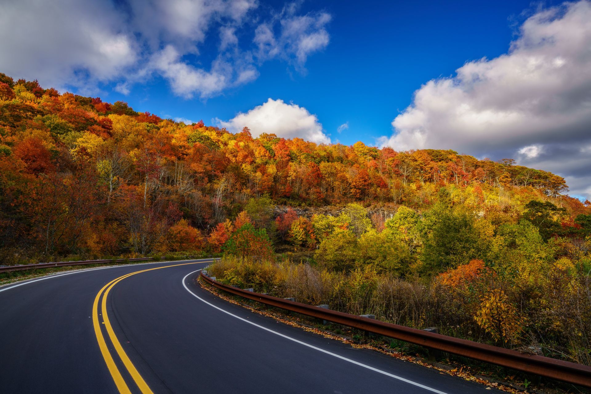 Winding road through colorful autumn trees under a bright blue sky with clouds.