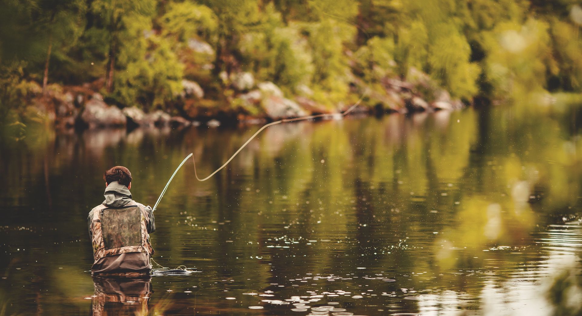 Person fly fishing in a calm river, surrounded by trees. The water reflects the forest and sky.