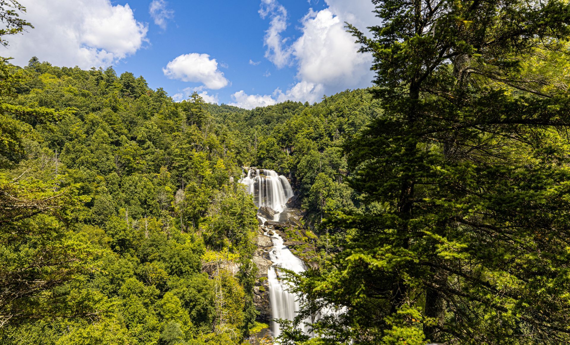 Waterfall cascading through lush green forest under a blue, cloudy sky.