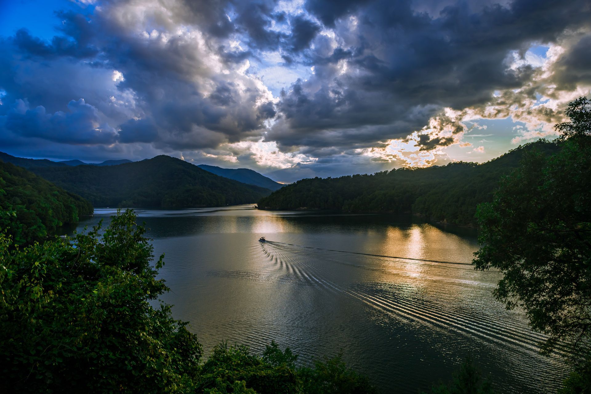 Dark clouds over a lake surrounded by mountains, sunlight breaks through. A boat leaves a wake.
