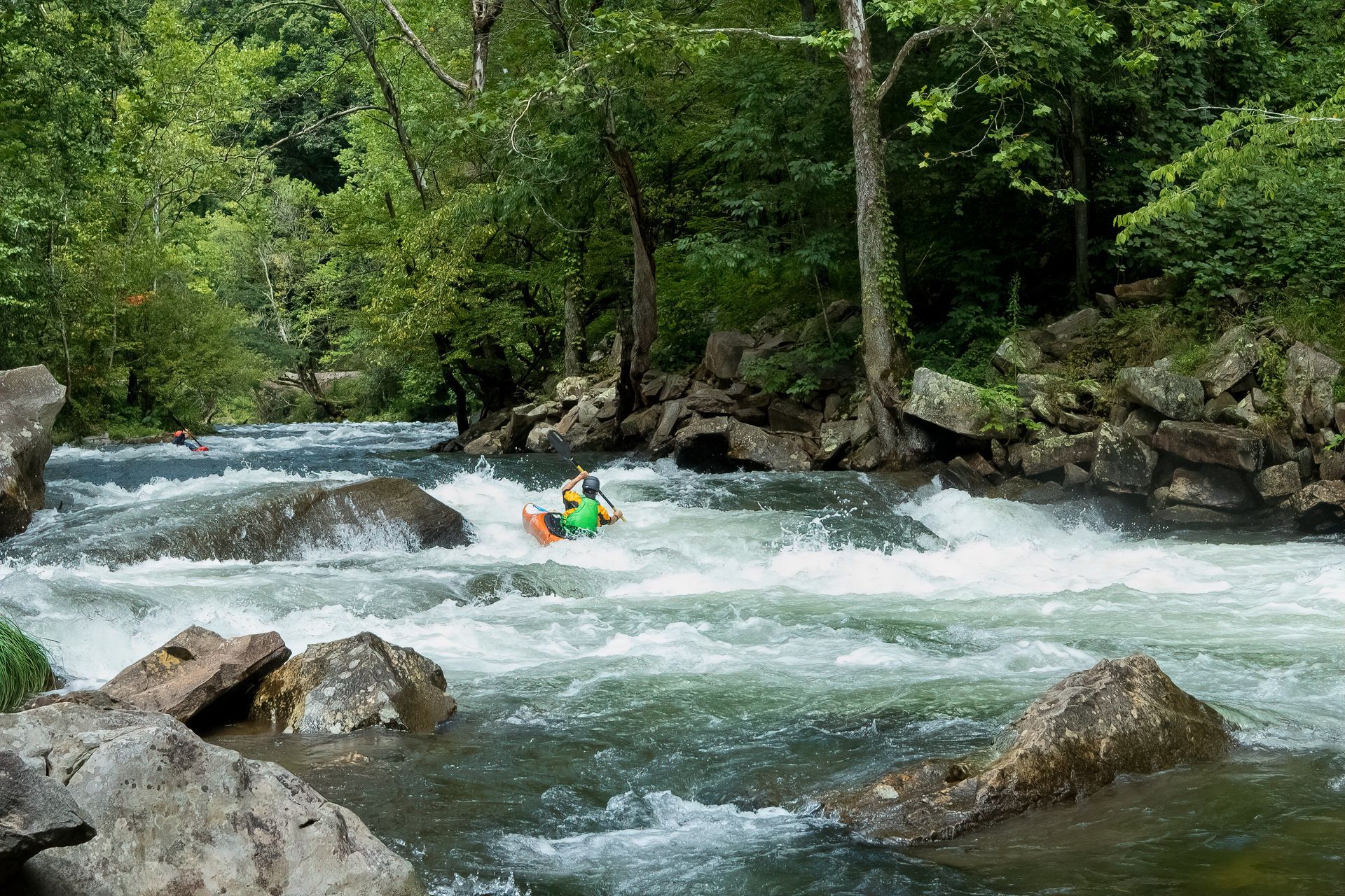 Kayaker navigates whitewater rapids, surrounded by rocks and lush green trees.