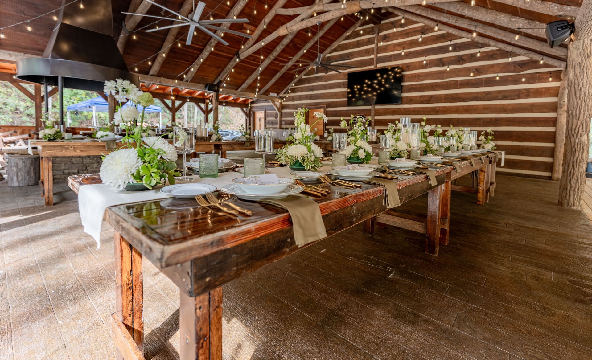 Long wooden table set for a formal gathering under a log cabin-style roof, decorated with white flowers and candles.