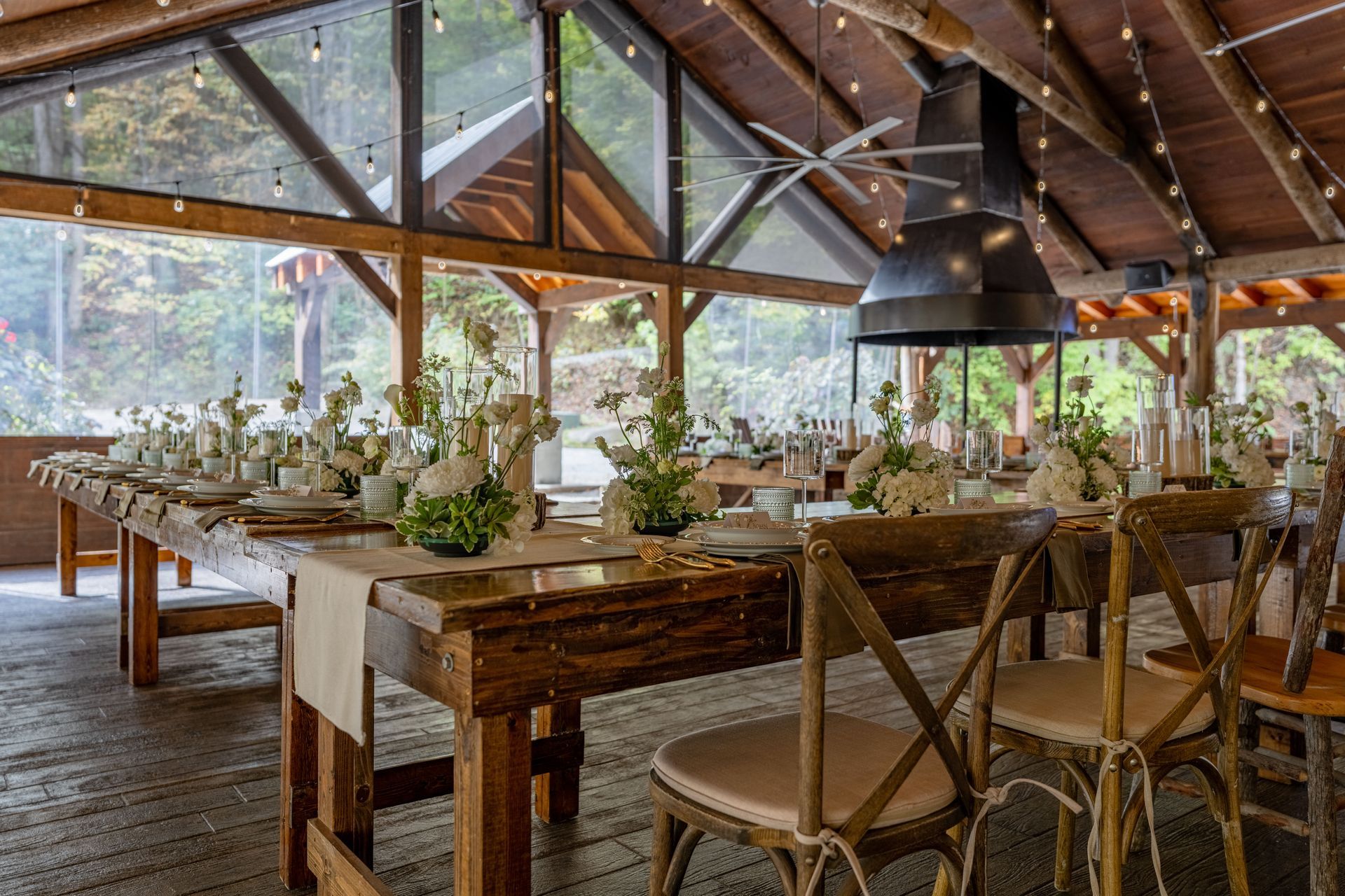 Long wooden tables set for a celebration in a rustic, open-air pavilion. White floral centerpieces.