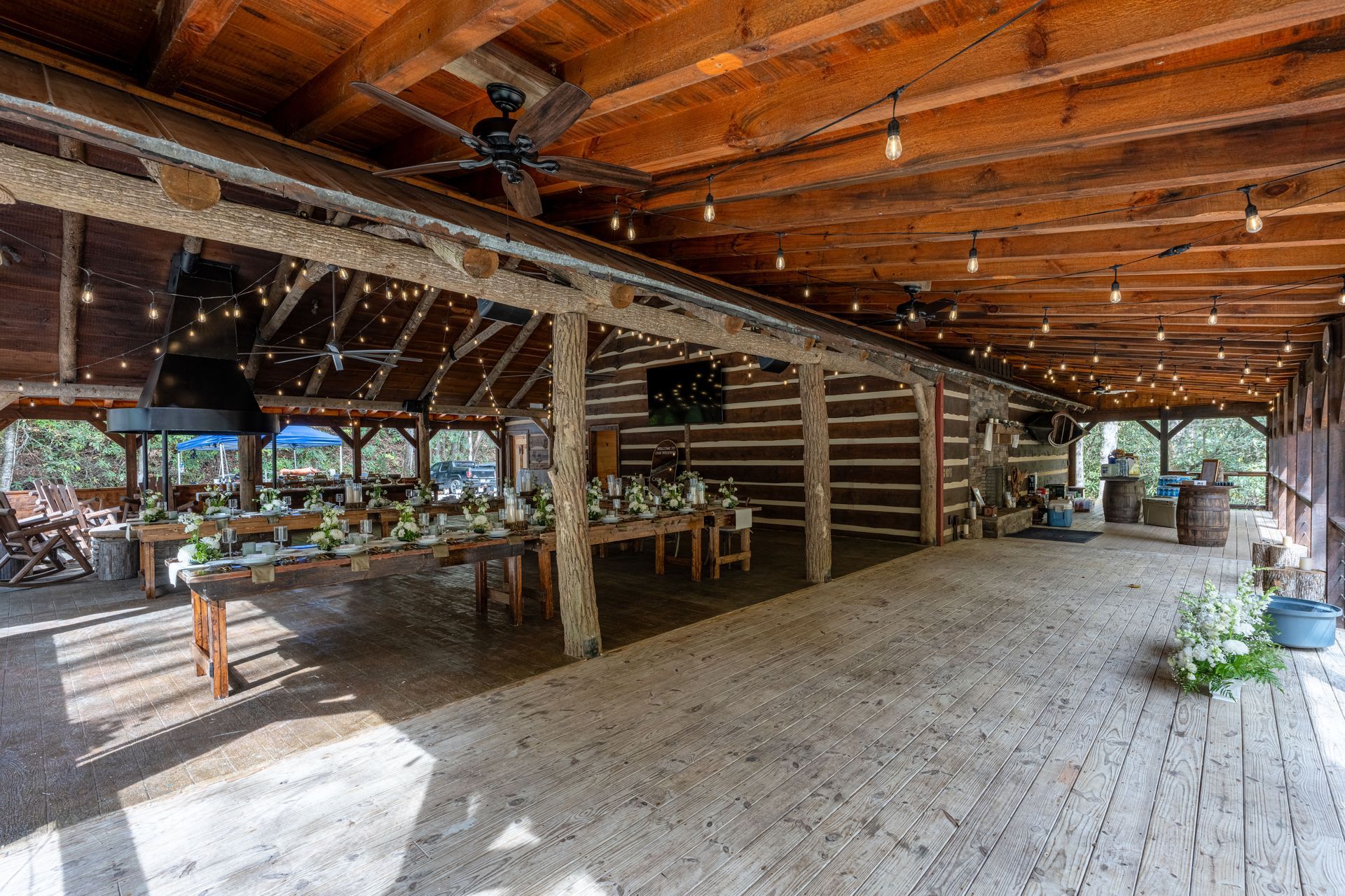 Rustic outdoor event space with wooden beams, log walls, string lights, and tables set for a gathering.