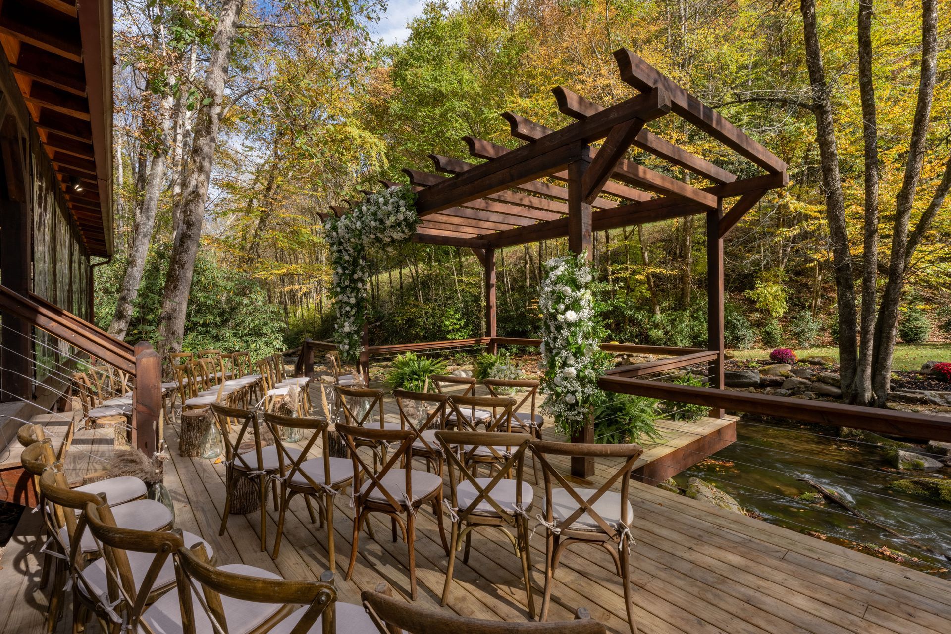 Outdoor wedding ceremony setting with wooden pergola and chairs near a stream. Autumn leaves.