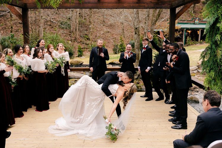 Bride and groom kiss, dipped on a wooden bridge, wedding party cheers in a forest setting.
