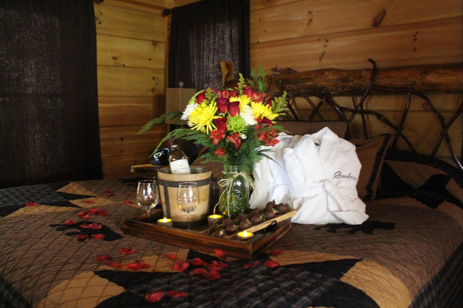 Romantic cabin bedroom with flowers, wine, chocolates, and robes on the bed.