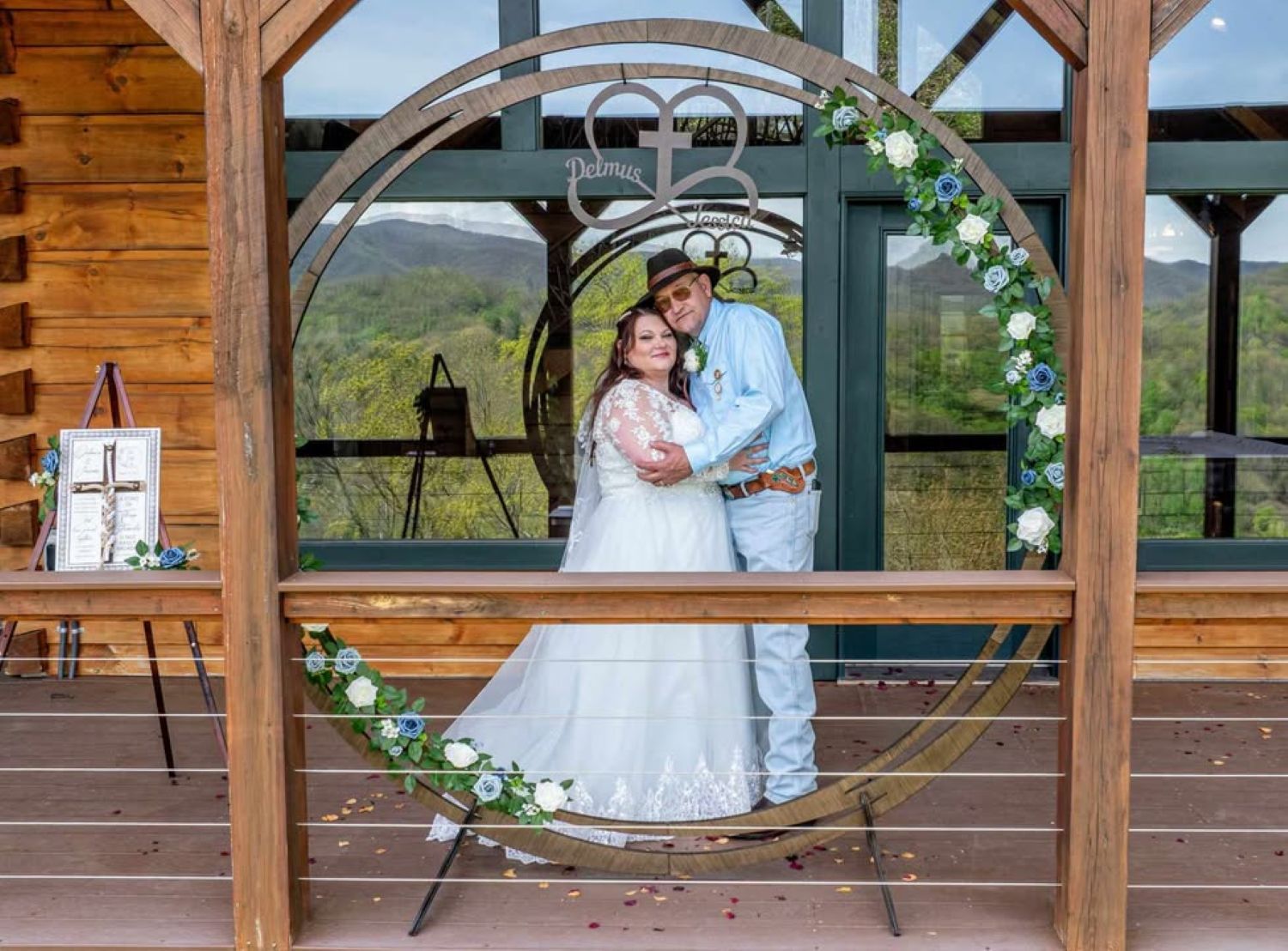 Couple in wedding attire embrace, framed by a floral arch, overlooking a scenic mountain view.