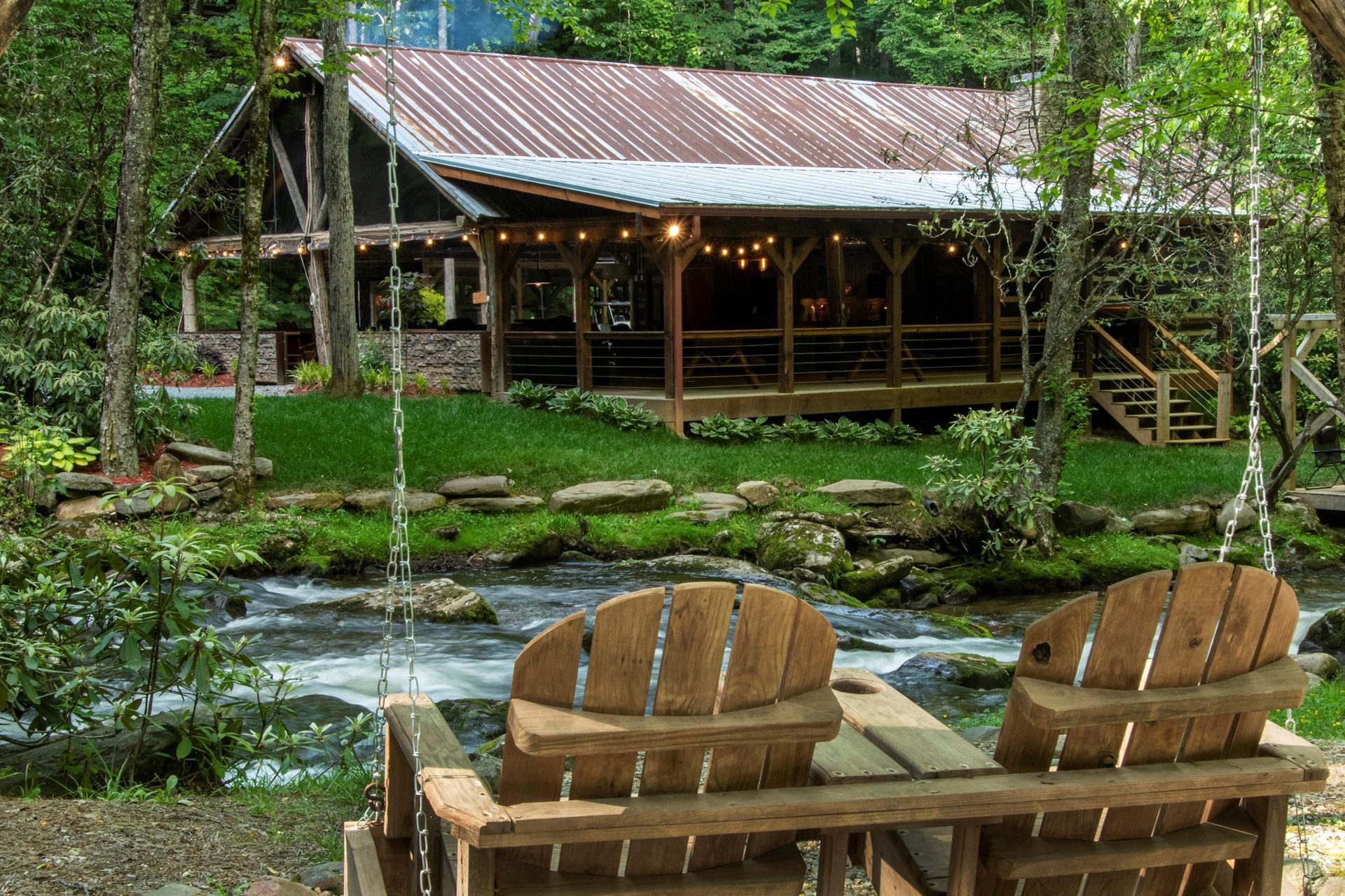 Cabin with porch and string lights next to a flowing river, surrounded by trees.