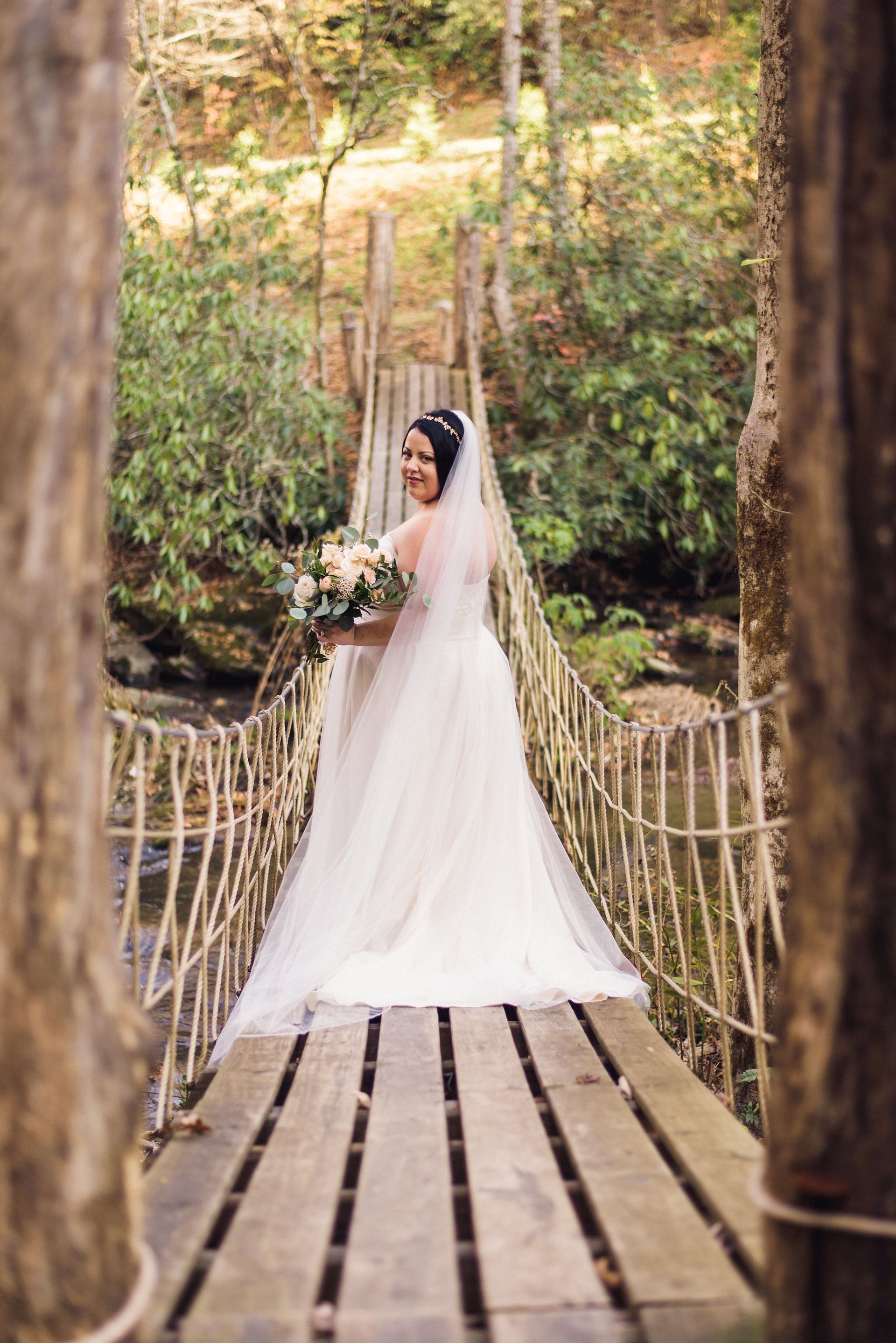 Bride in a white gown stands on a wooden suspension bridge, holding a bouquet, smiling. Forest background.