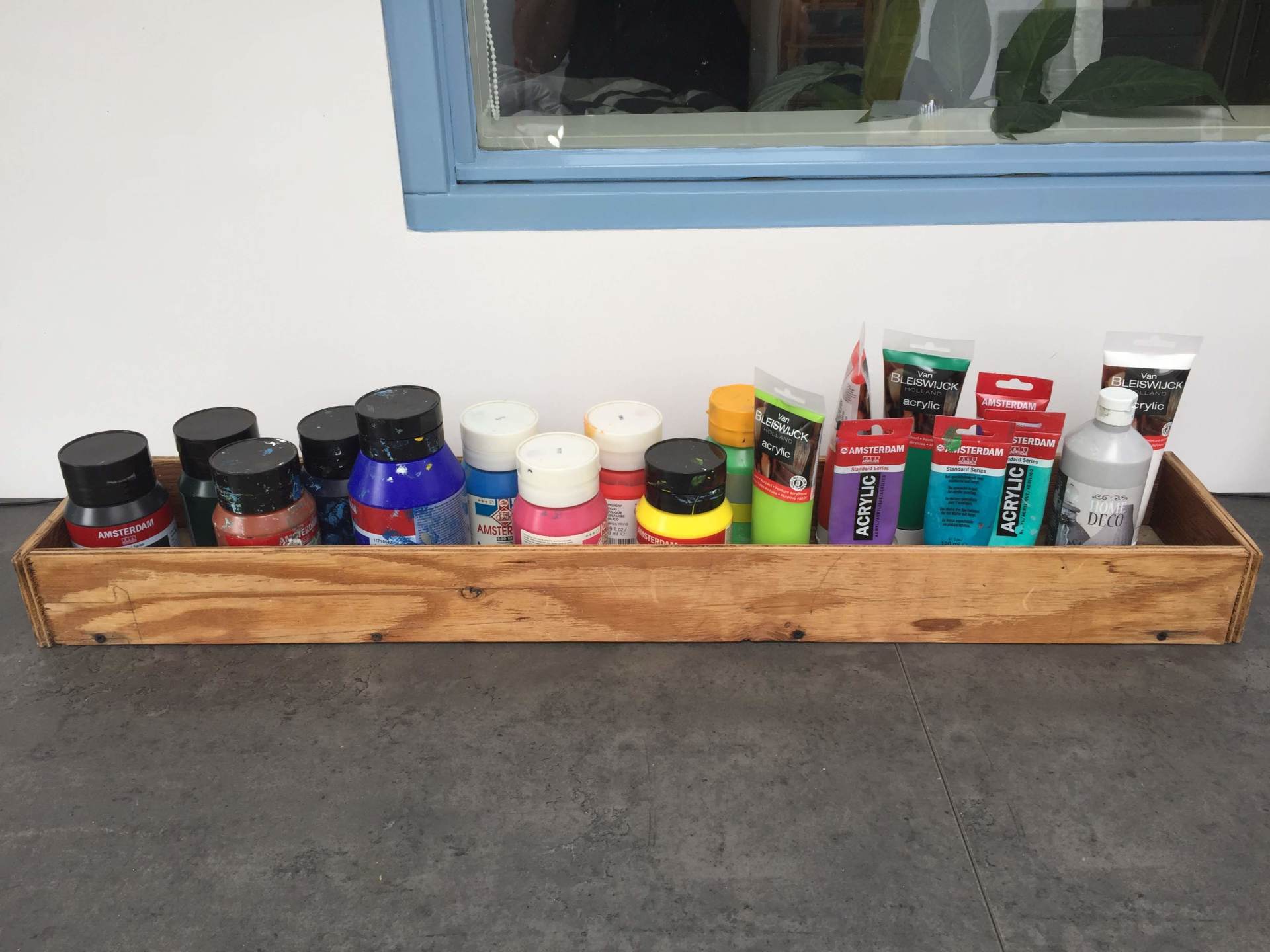Een lange houten schaal op een grijze ondergrond met daarop een assortiment van verschillende flesjes en tubes acrylverf.