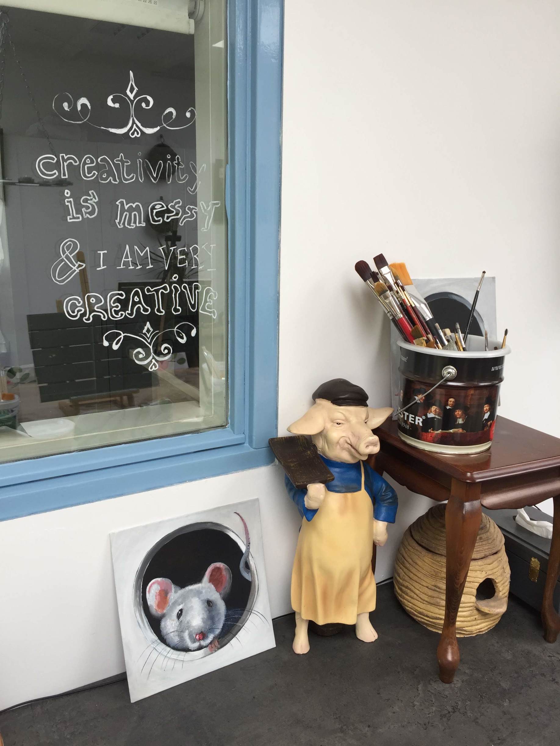 Een beeldje van een varkentje met een geel schortje staat naast een klein bijzettafeltje met kwasten, en op de vloer is een muizenpootafdruk te zien.