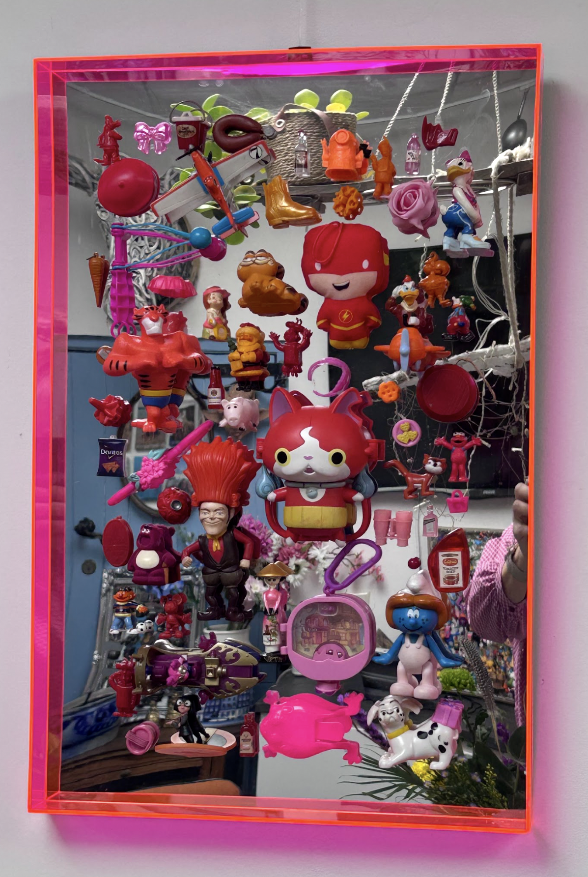 Een aan de muur bevestigde vitrine met een felroze neonframe, gevuld met een verzameling kleine, kleurrijke plastic speeltjes.