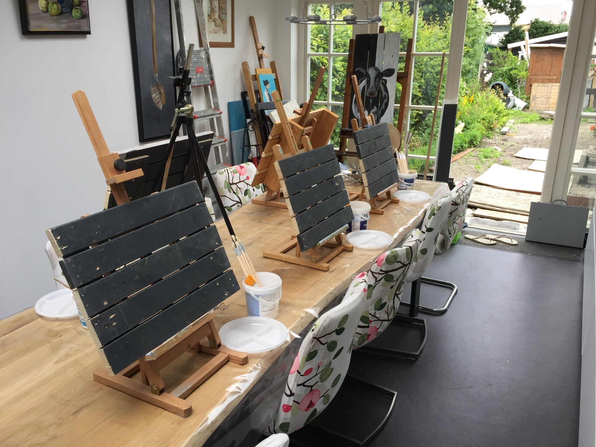 Een lichte kunststudio met een lange houten tafel, waarop meerdere schildersezels, doeken, verfbekers en stoelen met bloemenmotief staan.