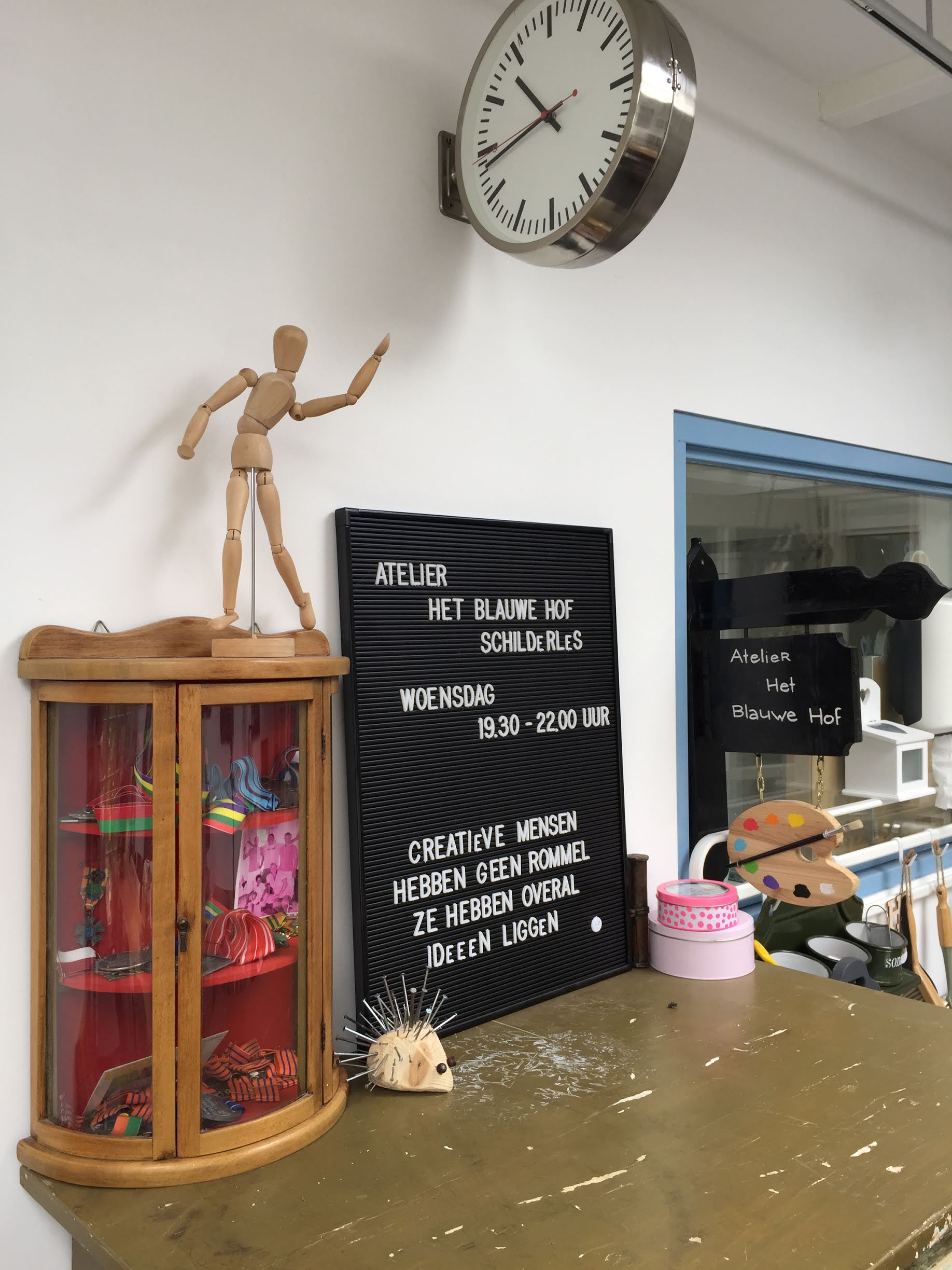 In een werkruimte staat een houten paspop op een glazen vitrinekast naast een bord en een wandklok.