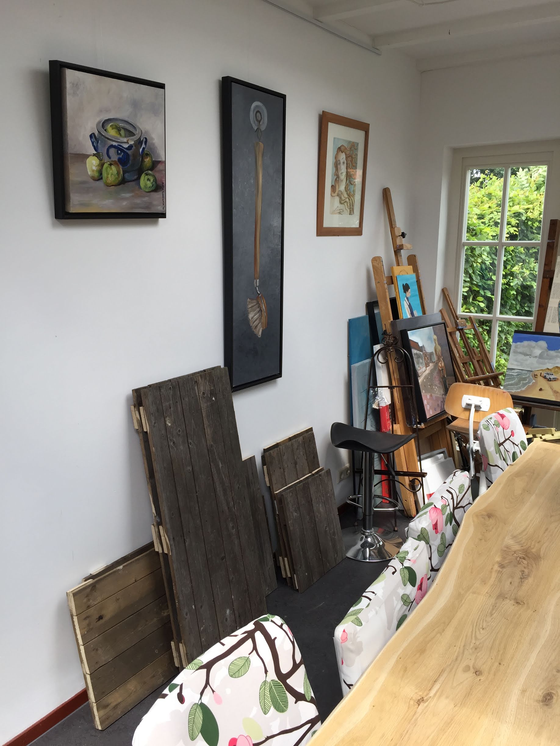 Een lichte kunststudio met schilderijen aan de muur, tegenaan leunende doeken, schildersezels en een houten tafel met stoelen met een patroon.