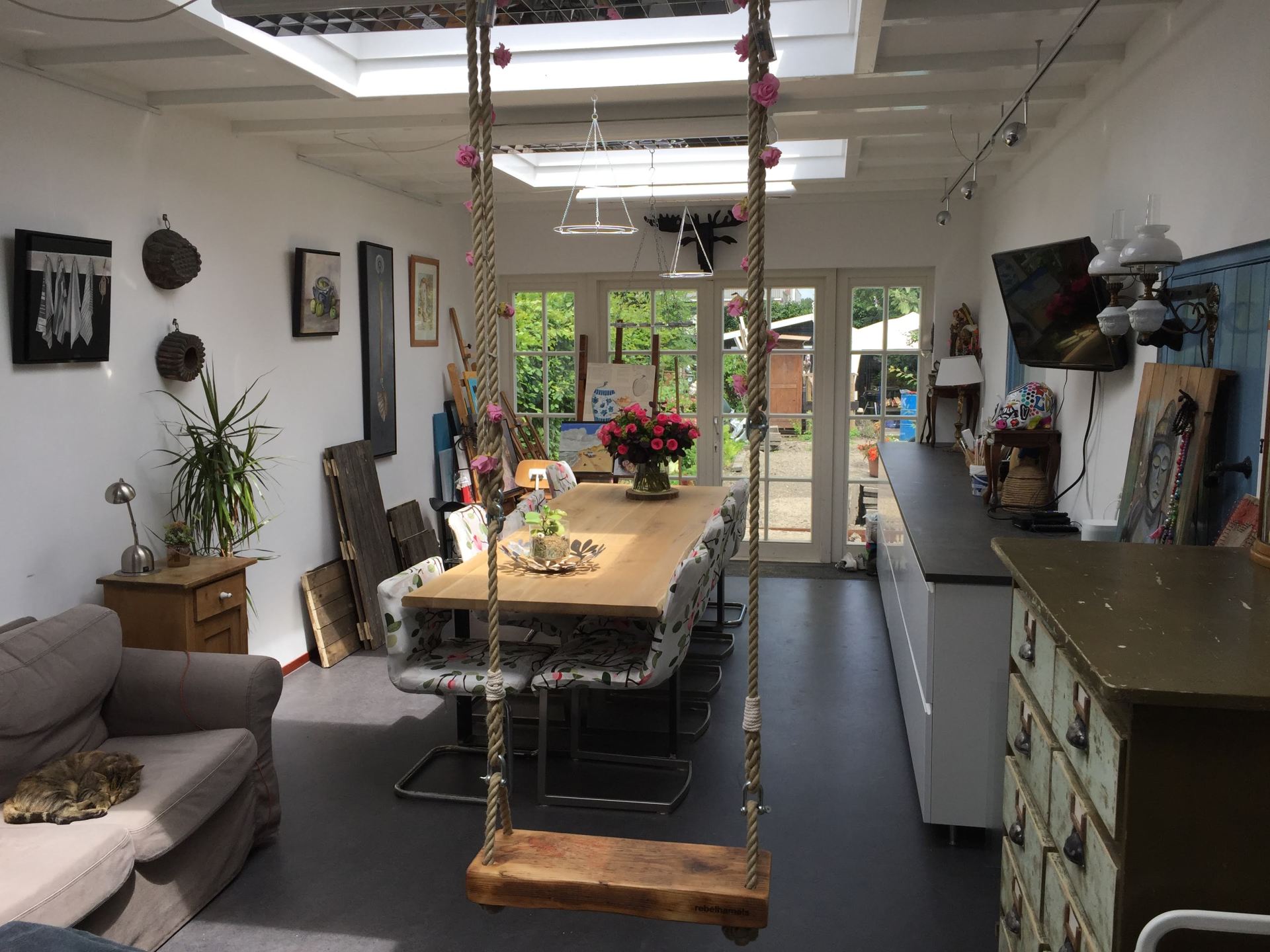 Een ruime, zonnige eetkamer is voorzien van een centrale houten eettafel, een hangende touwschommel en een bank aan de linkerkant.