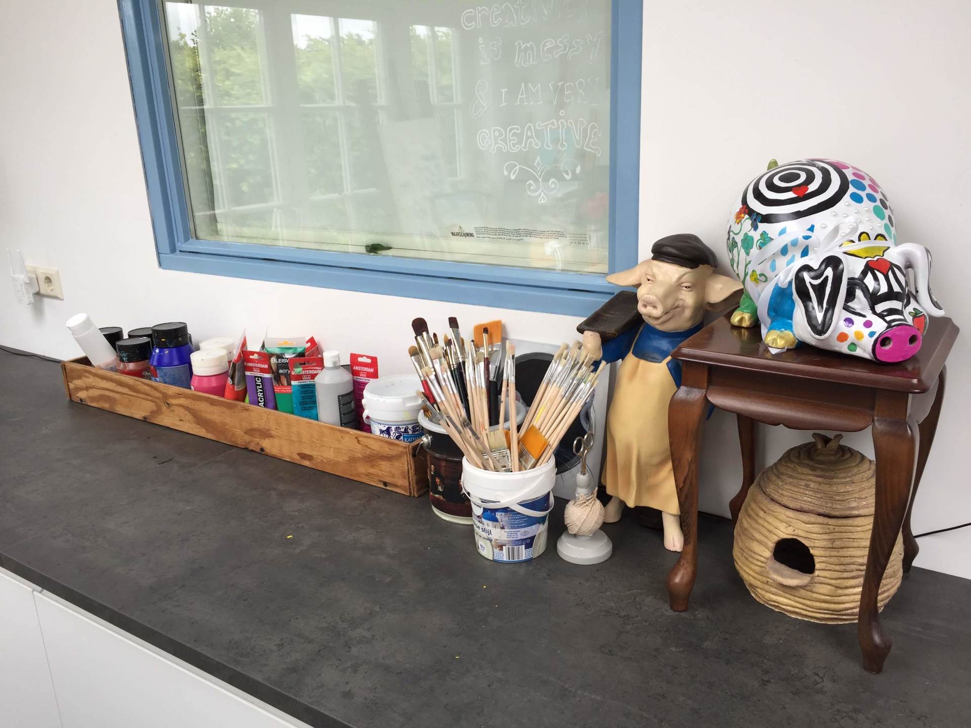 Tekenspullen op een houten dienblad, een kwastenhouder, een varkensbeeldje en een decoratief object op een klein tafeltje.