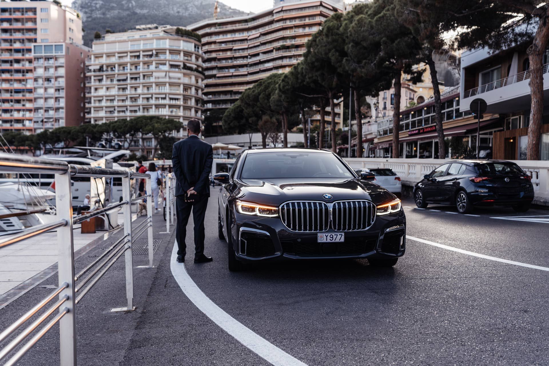 A man in a suit stands by a black BMW on a city street in front of buildings and the sea.