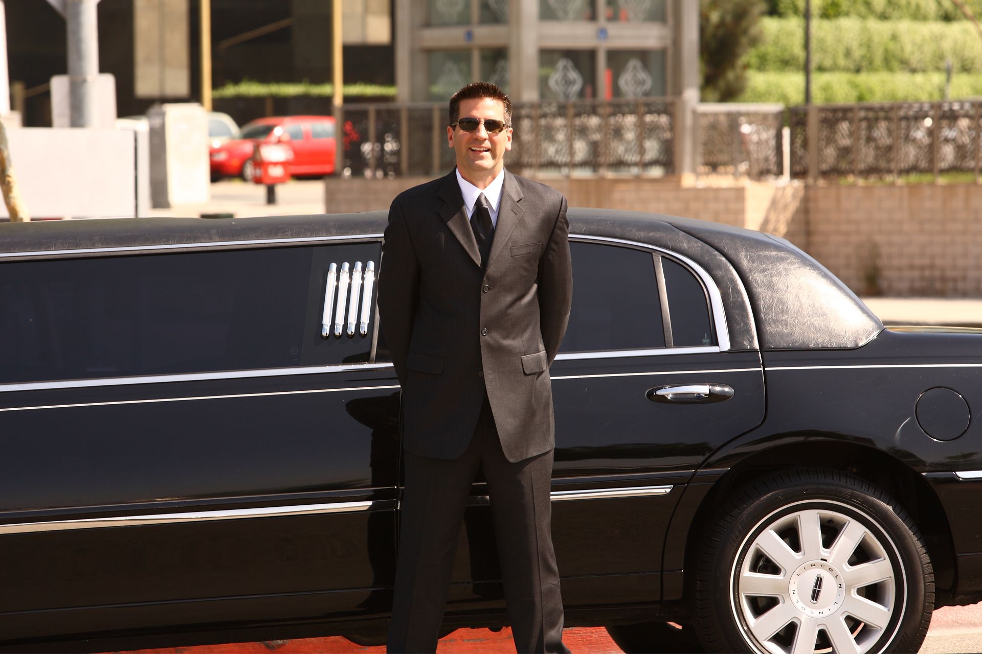 Man in a suit and sunglasses stands beside a black limousine.