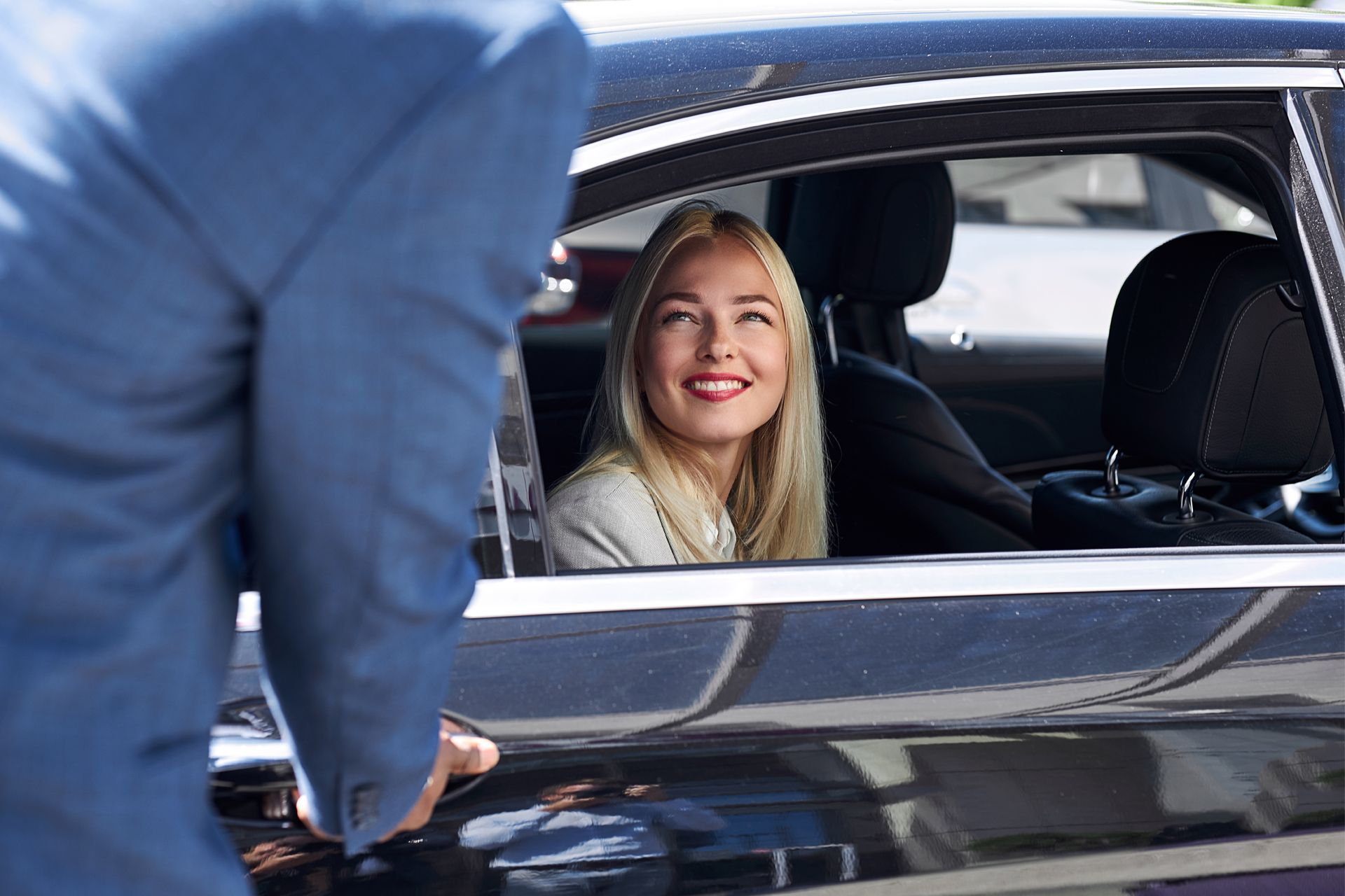 A woman with blonde hair smiles from a black car.