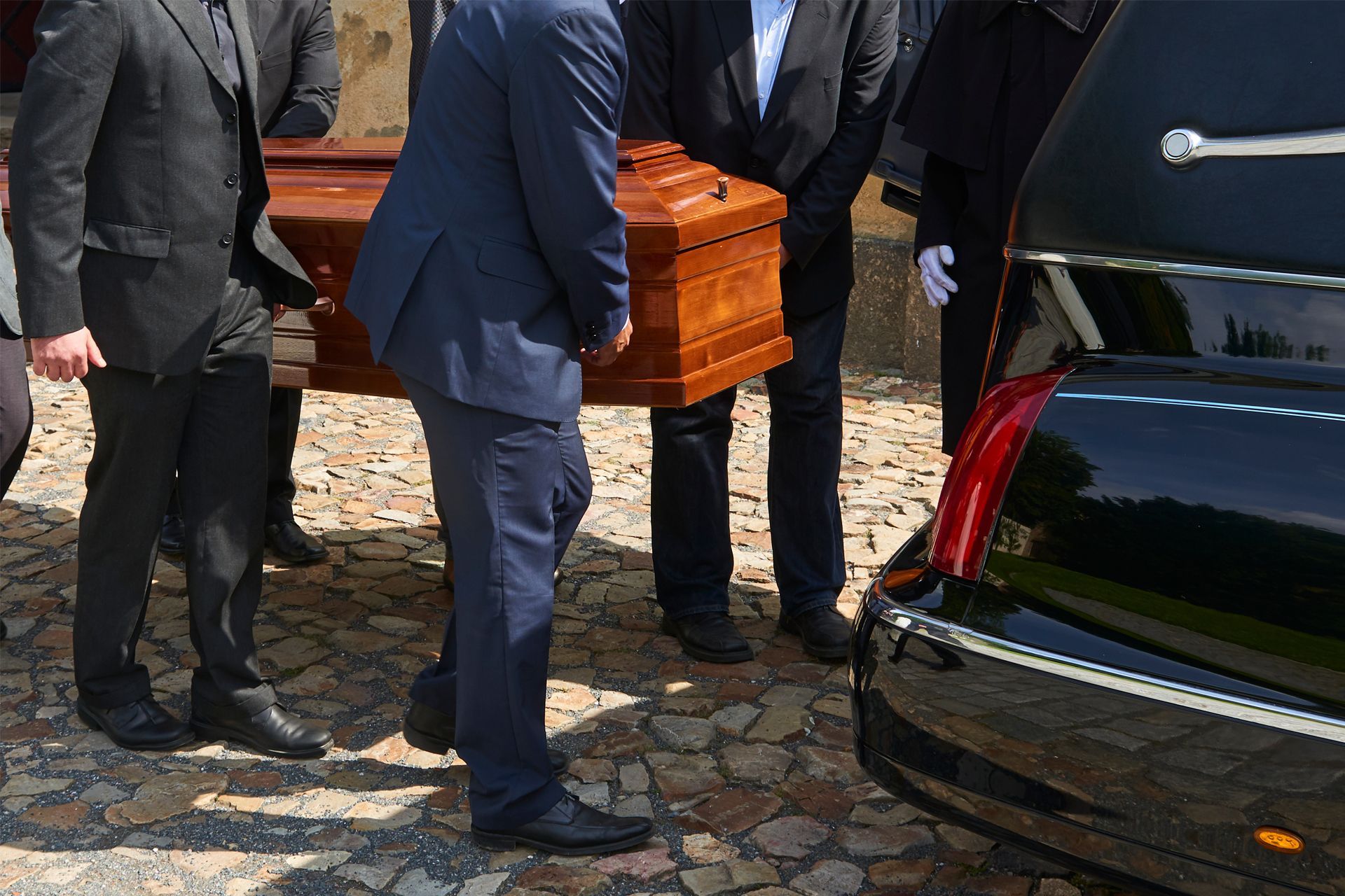 Men in suits carrying a wooden casket towards a black hearse on a cobblestone path.