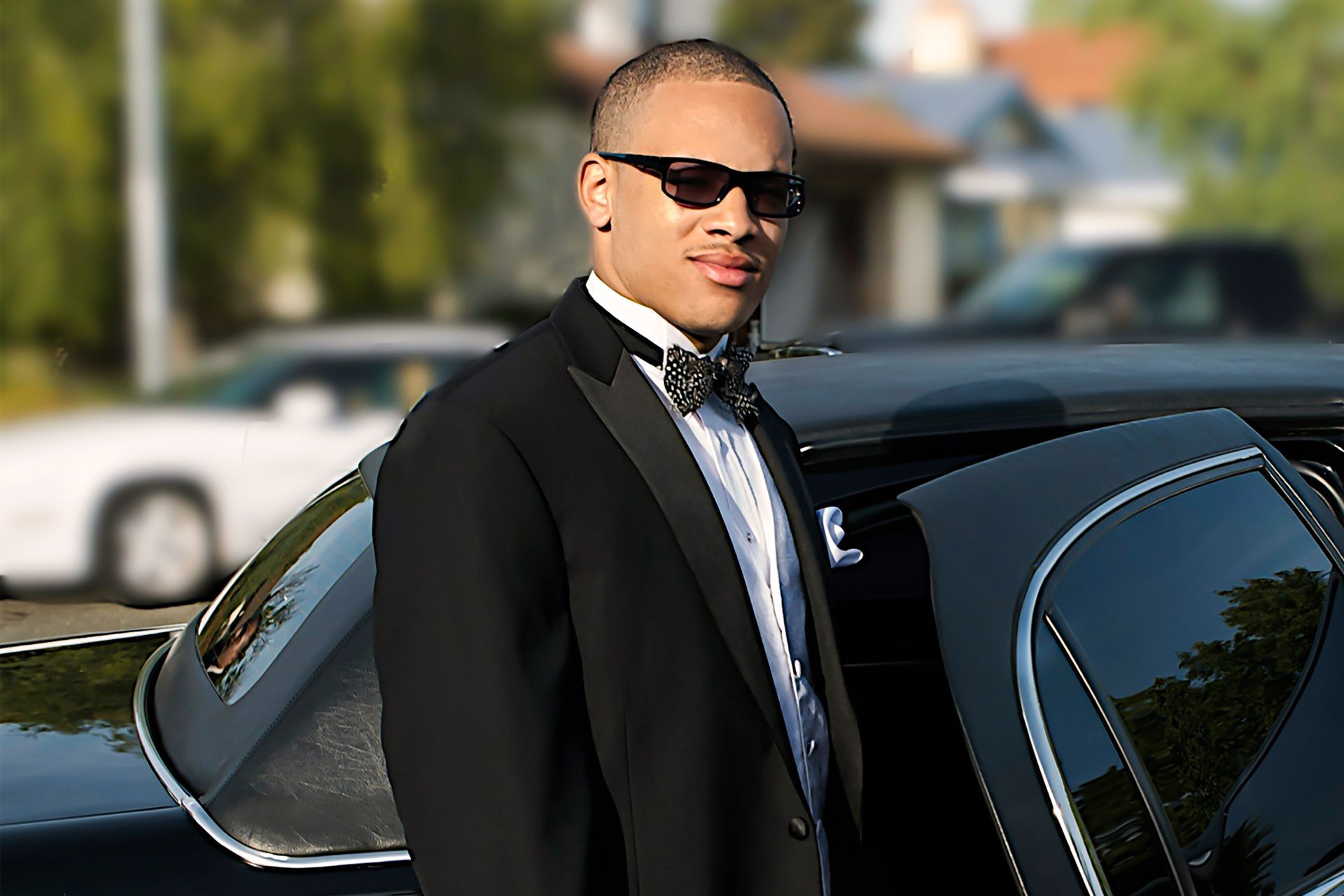 Man in a tuxedo and sunglasses standing by a black car likely prom night.