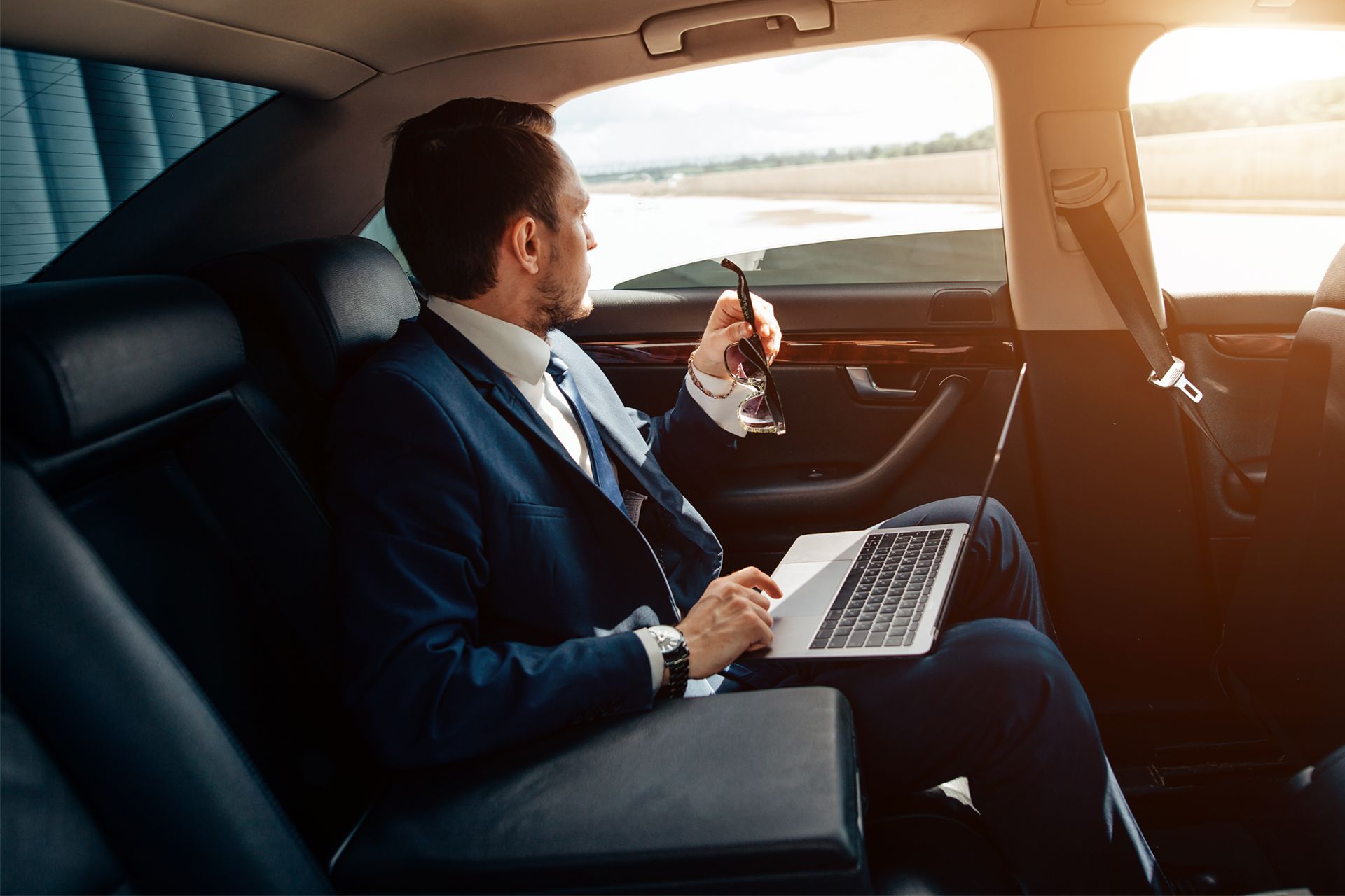 A businessman in a suit looks out the window of a car while holding a phone and laptop.