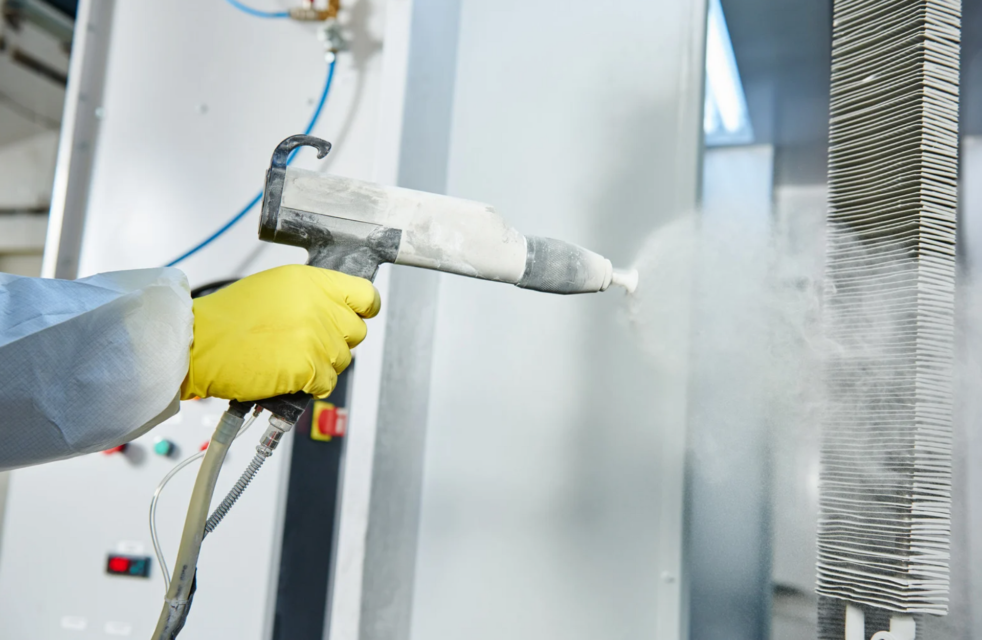 Person in yellow gloves uses a powder coating gun to spray metal object in a factory setting.