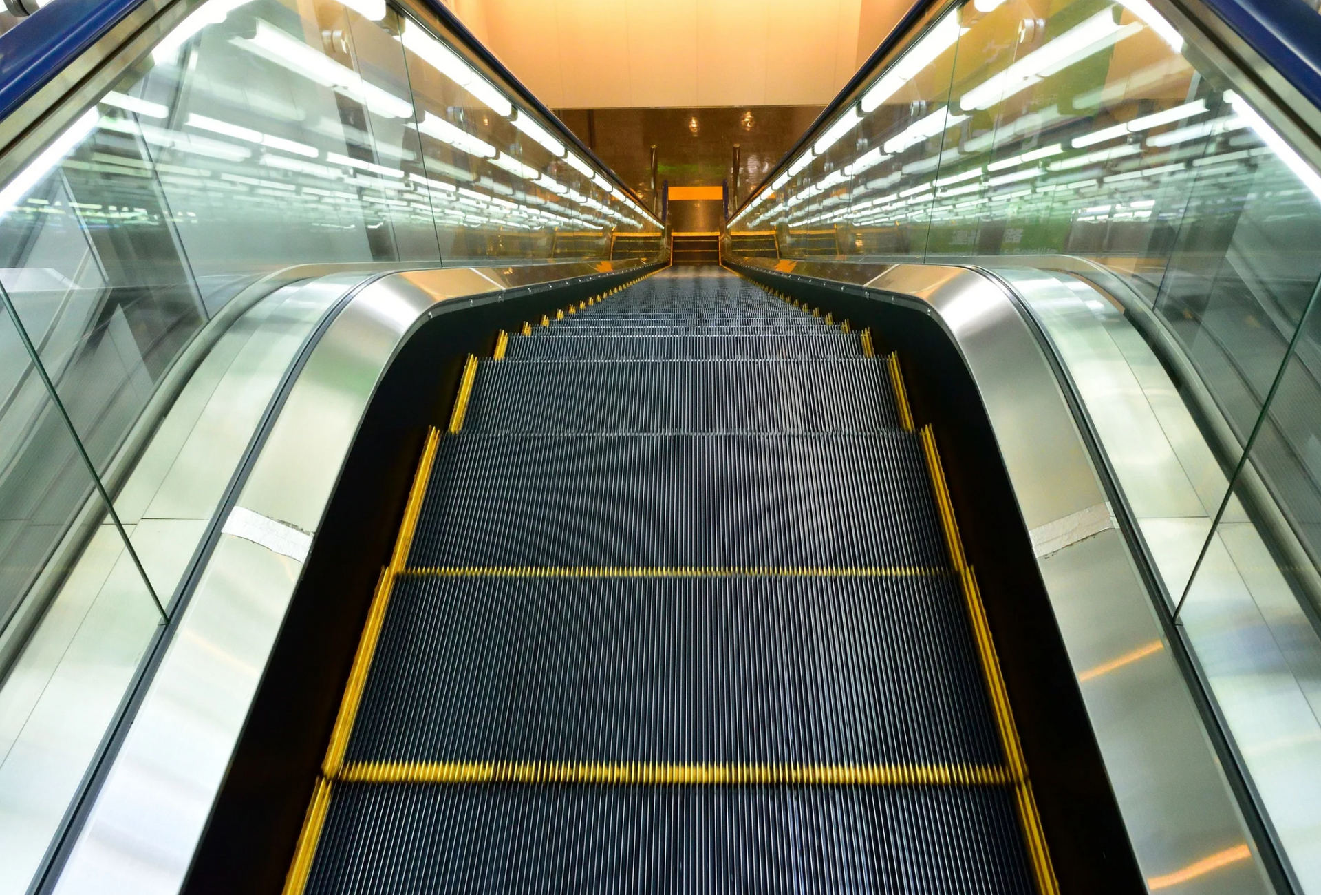 Escalator descending, stainless steel and glass sides, yellow safety lines, bright overhead lights.