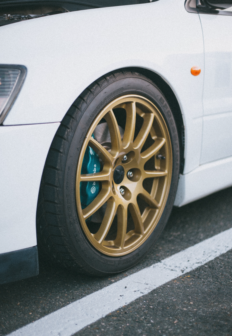 White car's front wheel with gold rim, teal brake, parked on asphalt, and a single white line.