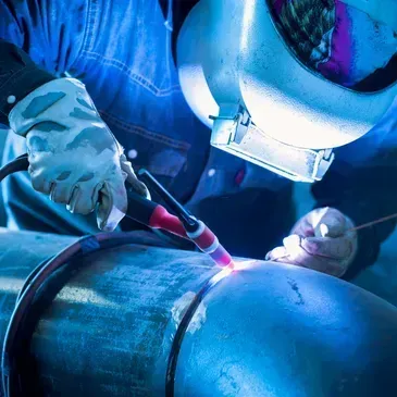 Welder in protective gear welding a large metal pipe, creating sparks and glowing light.