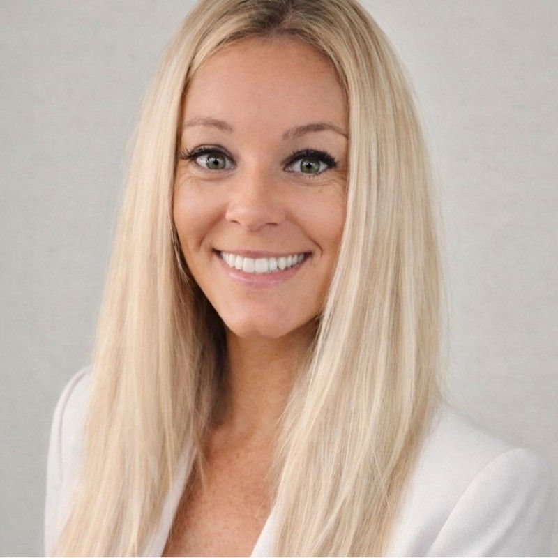 Blond woman in a white blazer smiling, headshot against a white background.