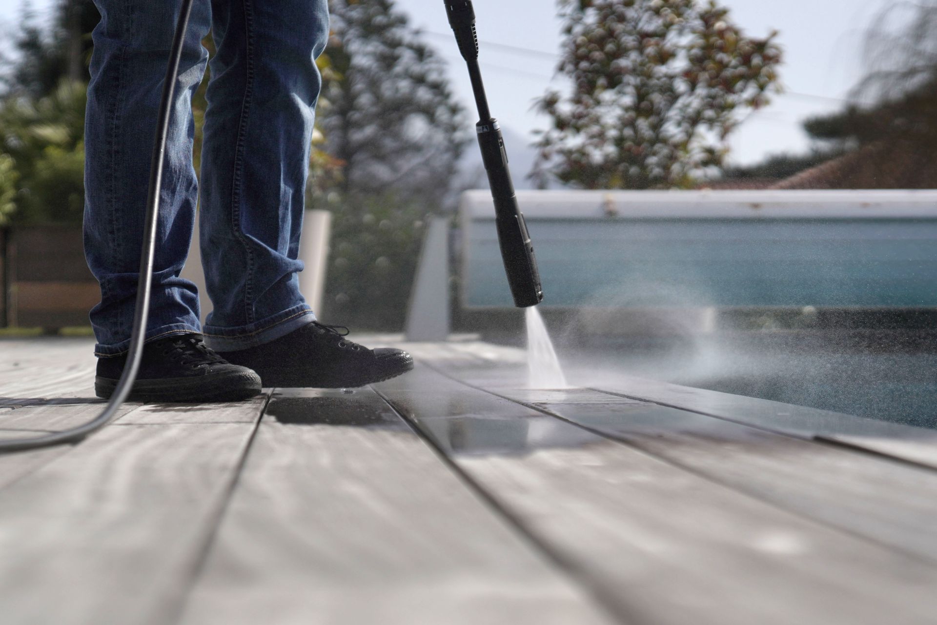 A person is using a high pressure washer to clean a wooden deck next to a pool.