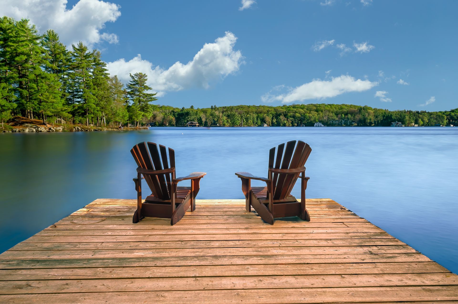 Two adirondack chairs are sitting on a wooden dock overlooking a lake.