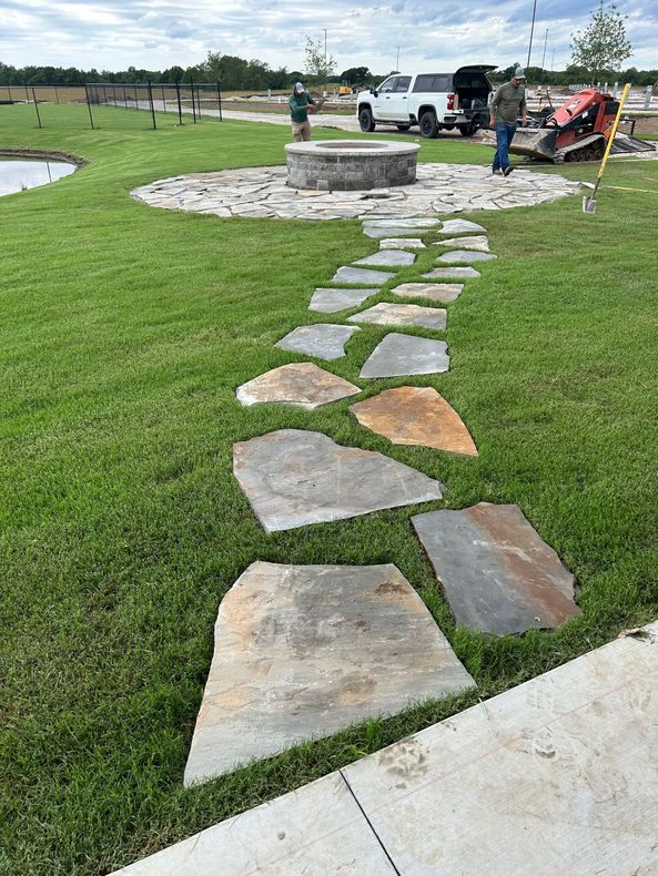 A stone walkway leading to a fire pit in the middle of a lush green field.