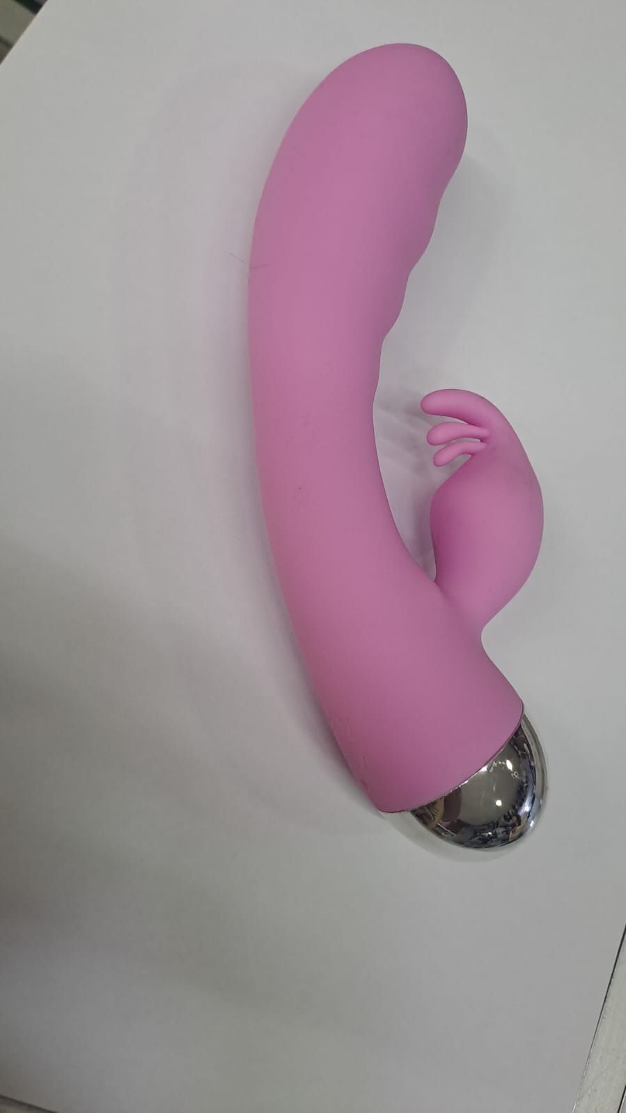 Vibrador rosa con eje curvado y apéndice estilo oreja de conejo, que reposa sobre una superficie blanca.