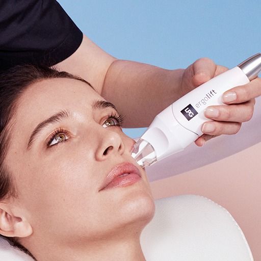 Woman receiving an Ergolift facial treatment. A handheld device is used on her face against a blue background.