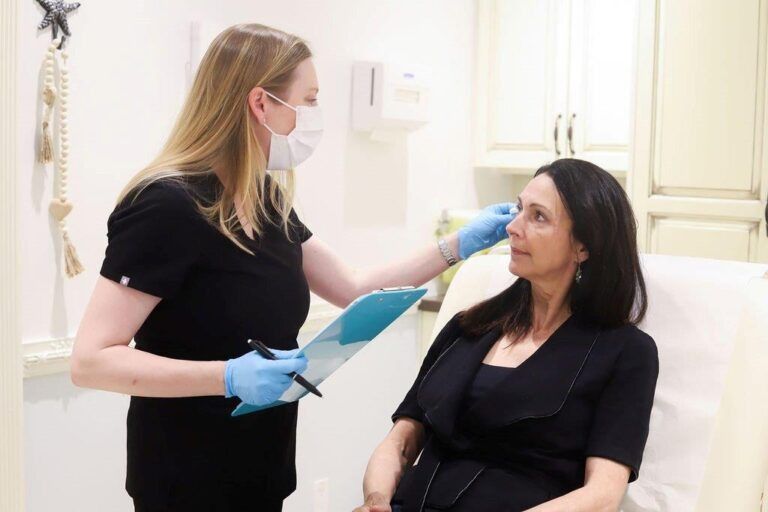 A medical professional, wearing a mask and gloves, examines a patient's face, holding a clipboard.