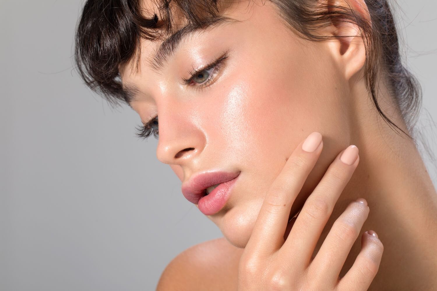 Woman with dewy skin, lightly touching her cheek. Soft lighting, pale pink lips.