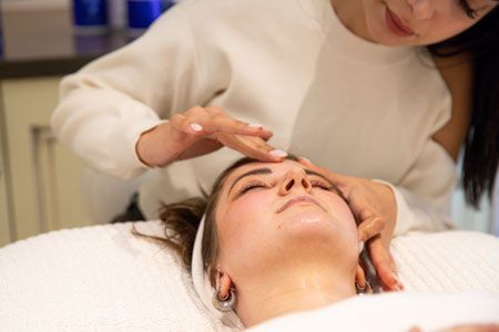 Woman receiving a facial treatment. Another person's hands gently touch her forehead. White sweater, bright setting.
